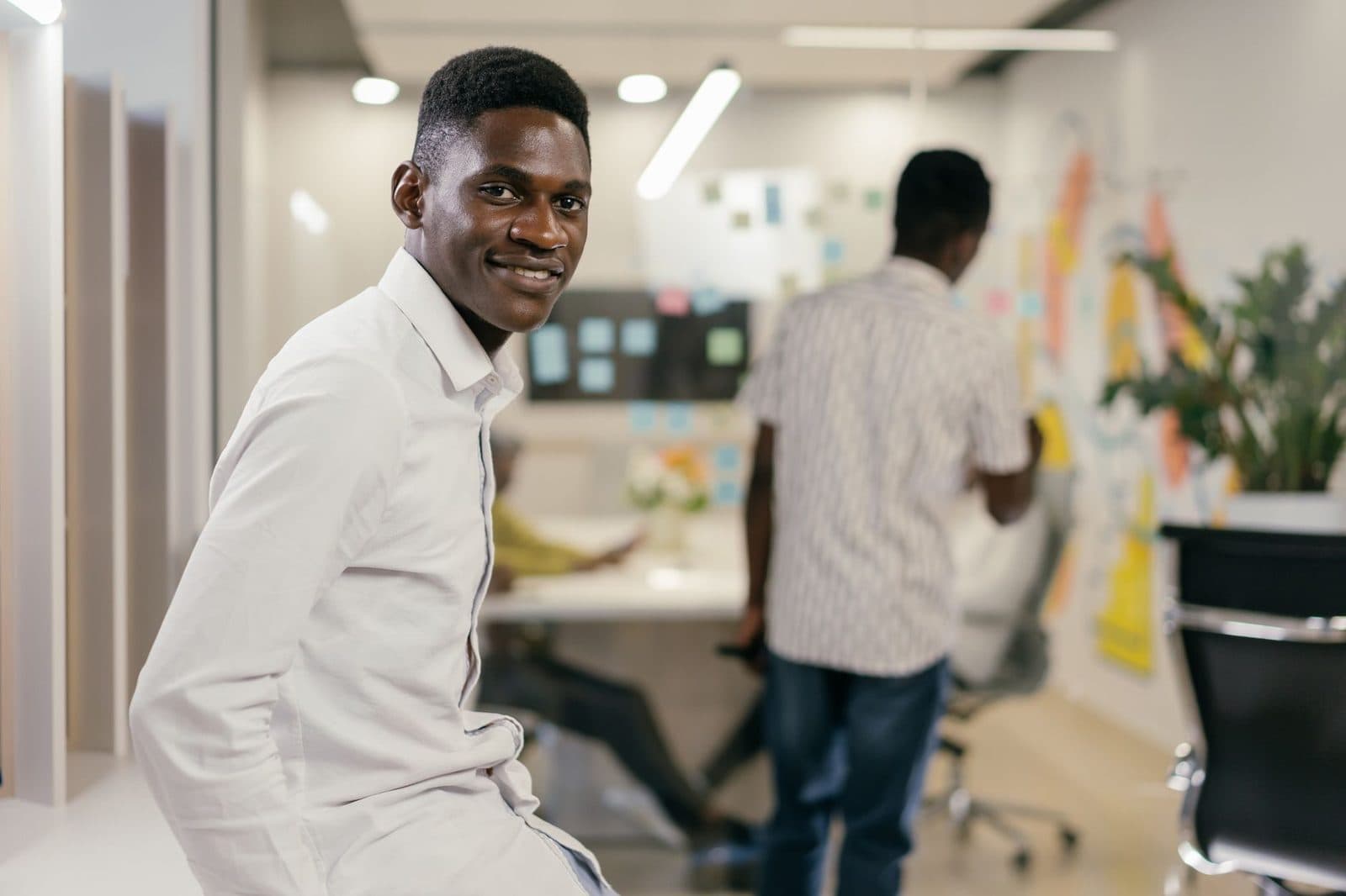 Jeune homme en chemise blanche souriant dans un bureau open space, collègues travaillant en arrière-plan
